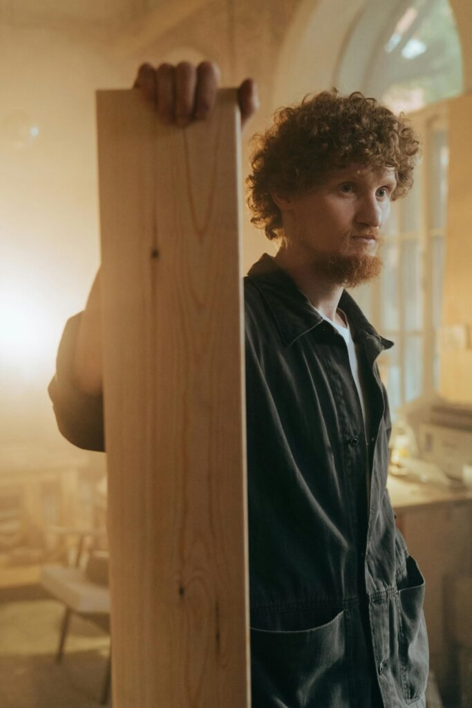 A skilled craftsman standing indoors, holding a wooden plank in a warm-lit workshop.